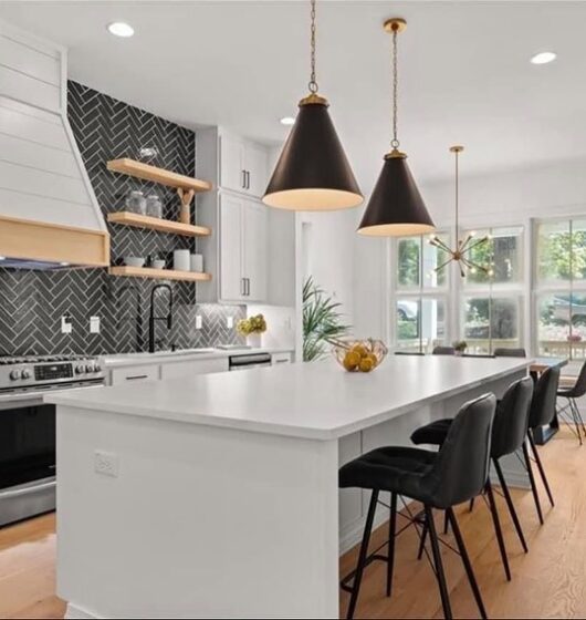 A modern kitchen with a white island, four black chairs, and two pendant lights. The backsplash is black with a herringbone pattern. White cabinetry, a stove, and open wooden shelves complete the look. A window seat with cushions is visible by the window.