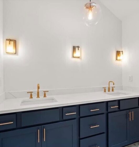 Modern bathroom with dark blue double vanity featuring gold hardware and fixtures. Two white sinks under brass light fixtures are mounted on a white wall. A glass pendant light hangs above, and a window to the left lets in natural light.