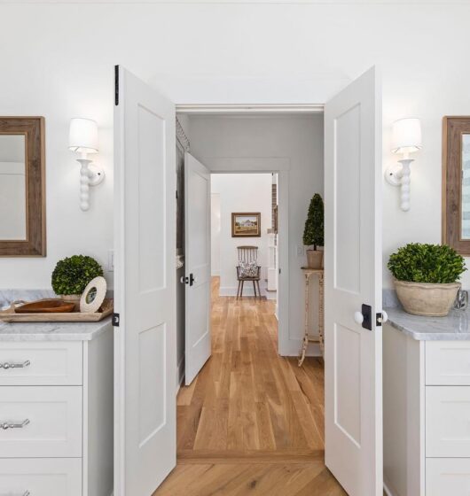 A bright bathroom features double vanities with marble countertops, two mirrors with wooden frames, and wall-mounted sconces. Doors open to a hallway with light wood flooring, and a glimpse of a closet on the left.