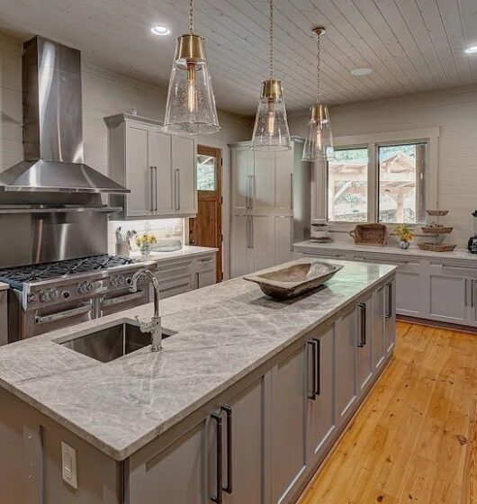 Modern kitchen with gray cabinets, a large marble island, and stainless steel appliances. Pendant lights hang above the island. The room has wooden floors, a clock on the wall, and a window with a view of trees.