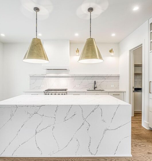 A modern kitchen with white marble countertops and island featuring gray veining. Two large gold pendant lights hang above the island. Stainless steel appliances are visible, with light wood flooring and a sliding glass door on the left.