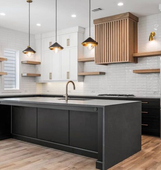 Modern kitchen with a large black island, white cabinets, and wood accents. Stainless steel fridge, built-in oven, and three pendant lights hang above the island. Light wood flooring complements the sleek design.