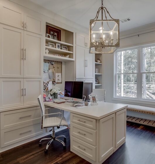 A home office with white built-in shelving and cabinets. A desk with a computer and chair is centered under a large window. The walls are decorated with photos and books, and a modern chandelier hangs from the ceiling. The flooring is dark wood.