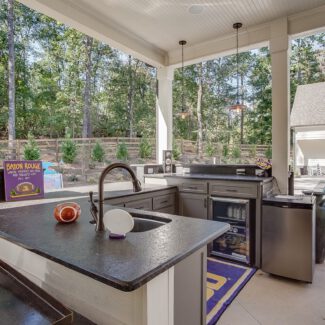 Outdoor kitchen overlooking a pool area with bar countertops, a sink, and a beverage cooler. Trees and a fence are in the background. A sign saying Baton Rouge leans against the counter, and a nearby house is visible across from the pool.