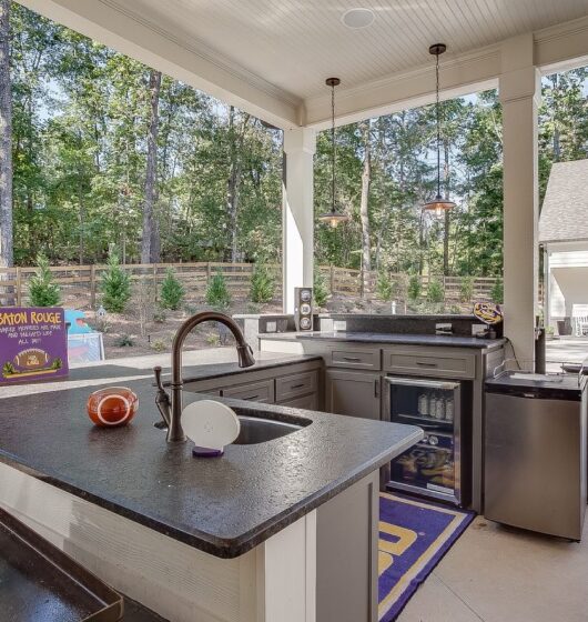 Outdoor kitchen overlooking a pool area with bar countertops, a sink, and a beverage cooler. Trees and a fence are in the background. A sign saying Baton Rouge leans against the counter, and a nearby house is visible across from the pool.