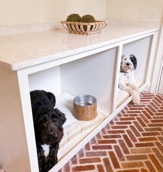 Two dogs are resting in built-in cubbies under a white marble countertop. Each cubby has a cushion and a wooden bowl. The floor is brick, and a basket of decorative green spheres is on the countertop.