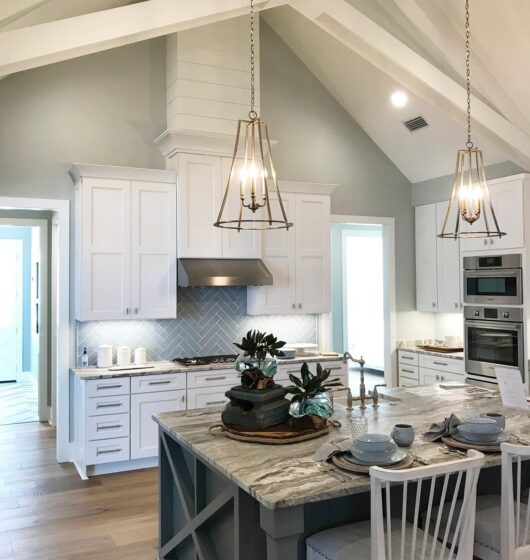 Bright kitchen with high ceilings, featuring white cabinets, a marble island with seating, and decorative plants. Two pendant lights hang above the island, and stainless steel appliances are built into the cabinetry. Light wood flooring complements the space.