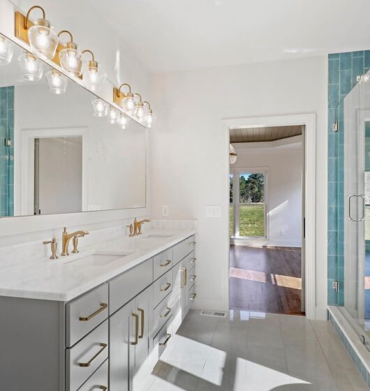 Bright modern bathroom with a large mirror, dual sinks, and pendant lights above a gray vanity. A glass-enclosed shower with blue tiles is on the right. An open doorway reveals a sunlit room with wooden flooring beyond.