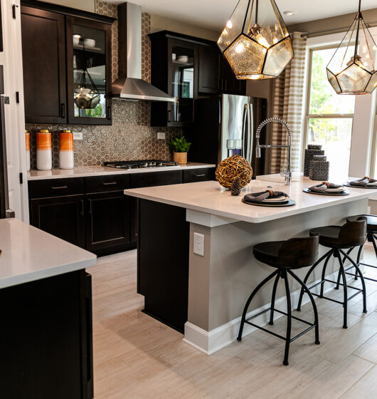 Modern kitchen with dark cabinets, stainless steel appliances, and light countertops. An island features three bar stools, decorative items, and pendant lights above. A large window with patterned curtains illuminates the space.