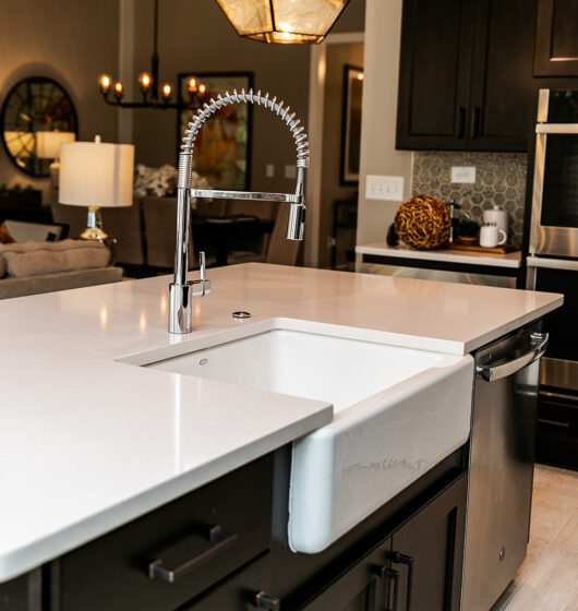 Modern kitchen interior featuring a sleek island with a white countertop and farmhouse sink. A chrome faucet is prominent. In the background, there are dark cabinets, a double oven, and a cozy living area with lamps and decorative mirrors.