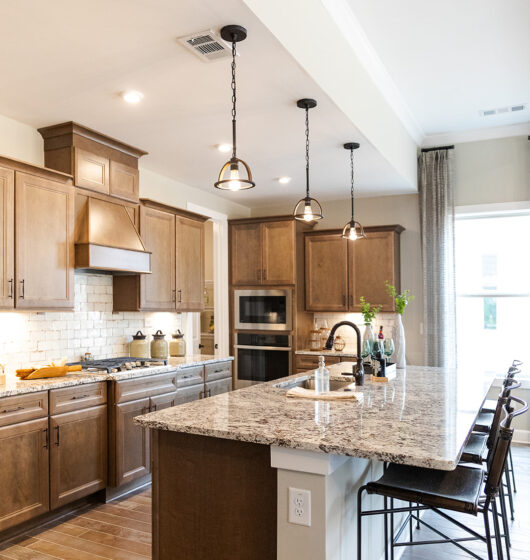 Modern kitchen with wooden cabinets, granite countertops, and stainless steel appliances. Features an island with bar stools, pendant lights above, and a window with curtains to the right. White subway tile backsplash adds a classic touch.