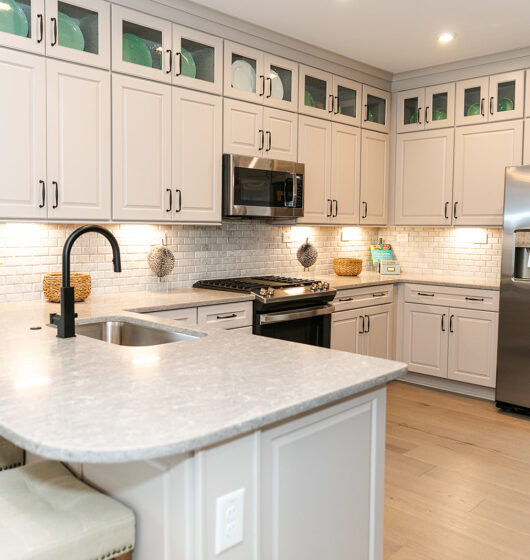 A modern kitchen featuring white cabinets, a stainless steel refrigerator, and a built-in microwave. The countertop is light gray with a black faucet. Green decorative plates are displayed above, and three stools line the breakfast bar.