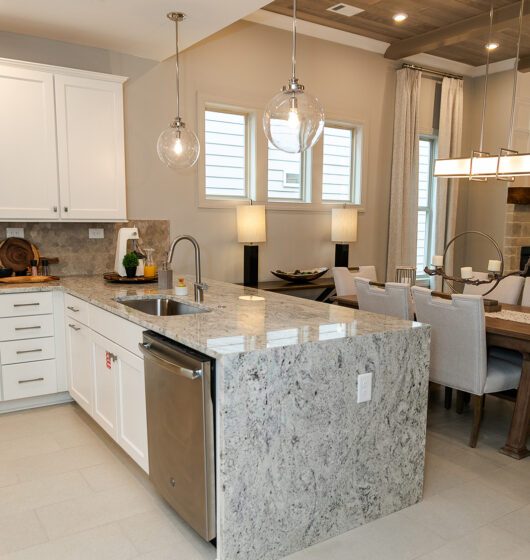 Modern kitchen with white cabinets and a gray marble island featuring a built-in dishwasher. Two clear pendant lights hang above the island. Adjacent is a dining area with a wooden table, upholstered chairs, and a rectangular chandelier.