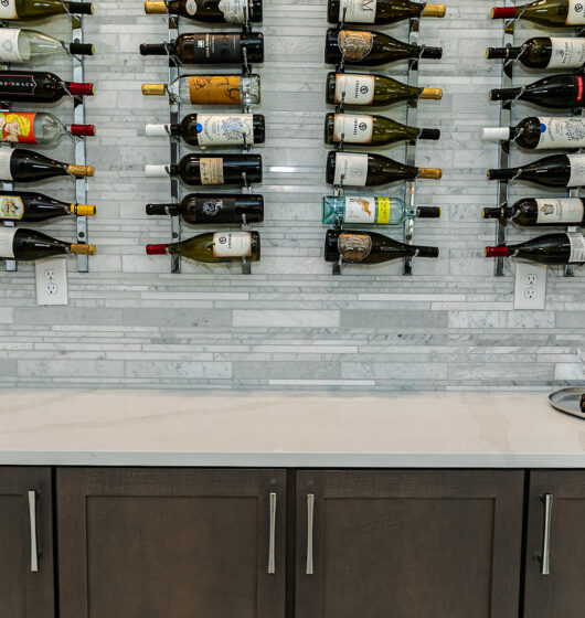 A wine display on a marble wall showcasing bottles organized on horizontal racks. Below is a white countertop with dark cabinets and a silver tray holding two empty wine glasses and a small pine cone. The scene is well-lit and elegant.