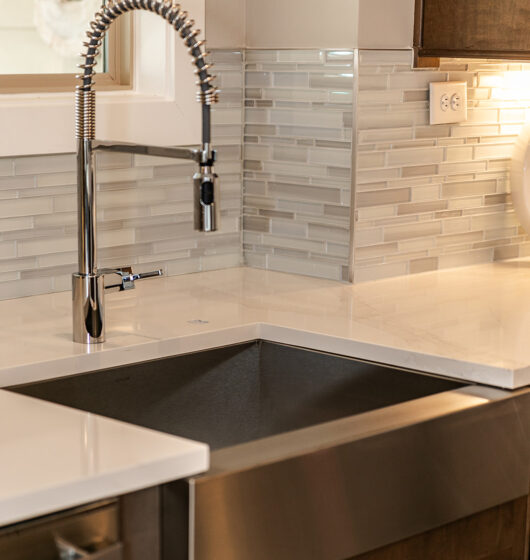 Modern kitchen sink with a high, flexible pull-down faucet. The backsplash is made of light gray elongated tiles. The countertop is white, and the cabinets are a dark wood finish. An electrical outlet is visible on the wall.