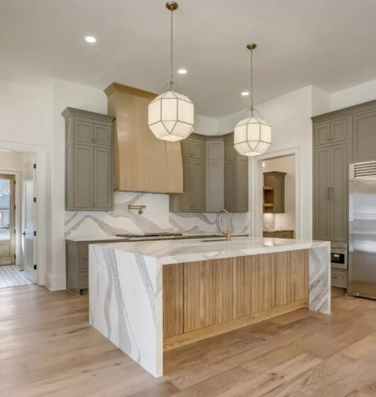 Modern kitchen with light wood flooring, marble countertops, and a large island featuring wooden accents. Gray cabinets and stainless steel appliances, including a double-door fridge. Two geometric pendant lights hang over the island.