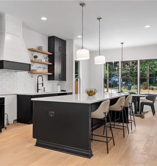 Modern kitchen with a large white island countertop, black base, four bar stools, and pendant lights. White hexagonal tile backsplash, black and wood cabinets, a black sink, and floating shelves. Adjacent dining area with large windows and a table.