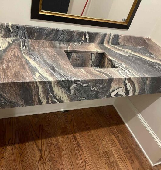 A bathroom sink with a rectangular basin is set into a marbled countertop featuring swirls of brown, black, and white. The sink is mounted against a white wall above a hardwood floor, with a rectangular mirror above it and a soap dispenser on the right.