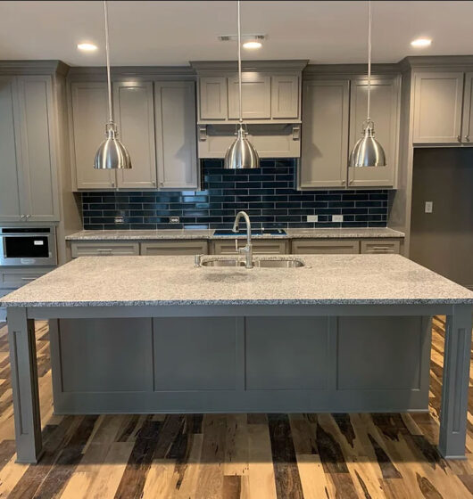 Modern kitchen with gray cabinets and a large island featuring a sink. Three pendant lights hang above the island. The backsplash is dark tiled, and the floor is wood. The kitchen is spacious and well-lit.