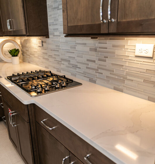 Modern kitchen with dark wood cabinets, a beige countertop, and a gas stove. A light gray subway tile backsplash lines the wall. Under-cabinet lighting illuminates the workspace. A small white potted plant decorates the counter.