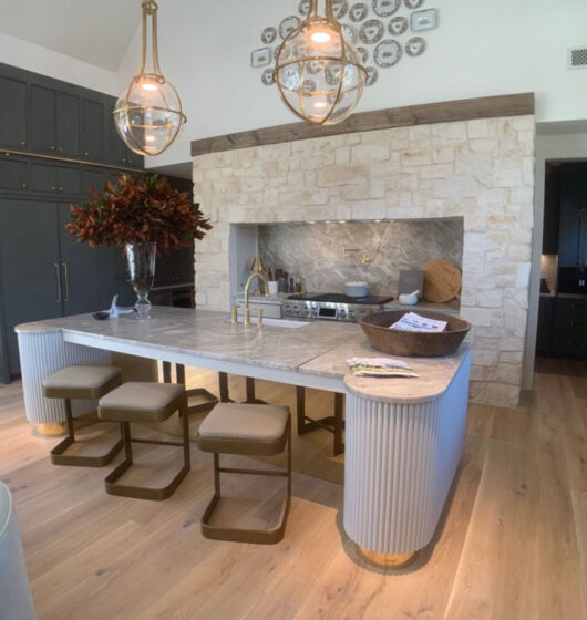 Modern kitchen with a stone backsplash and island with a marble top. Three bar stools are tucked under the island, which is adorned with two large pendant lights and a decorative plant. Light wood flooring and dark cabinetry complete the space.