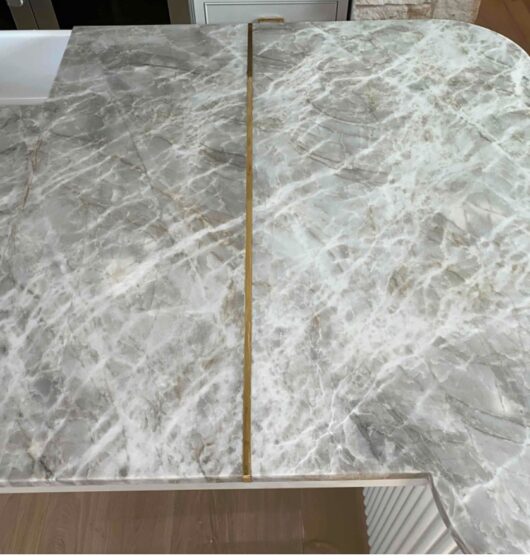 A marble countertop with a light gray and white veined pattern, featuring a visible seam with a gold strip running across it. The countertop edge is curved, and the surface appears smooth and polished.