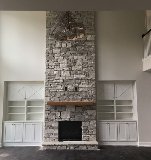 Spacious living room with high ceiling, featuring a tall stone fireplace centered between built-in shelves and cabinets. A wooden mantel decorates the fireplace. Natural light streams through large windows on the left side.