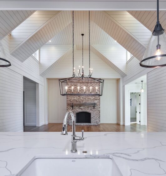 Luxurious kitchen view featuring a white marble countertop with a sink in the foreground. Hanging pendant lights, a brick fireplace, and a vaulted shiplap ceiling with exposed beams in the background create an elegant and spacious ambiance.