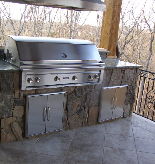 A stainless steel outdoor grill is built into a stone countertop on a patio. It has multiple burners and storage cabinets below. The area overlooks a natural, wooded landscape with bare trees, suggesting a winter setting.