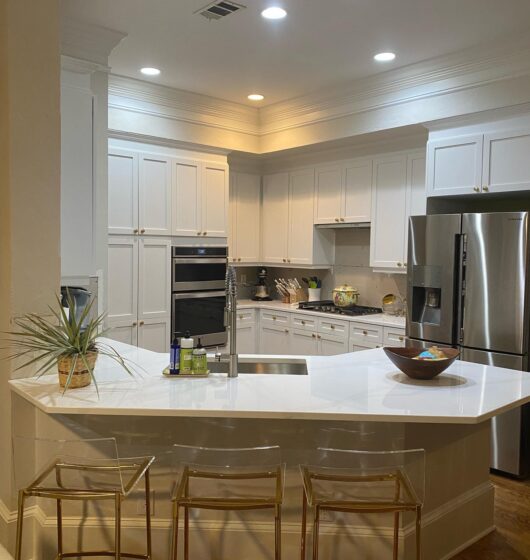 Modern kitchen with white cabinets and stainless steel appliances. A white island features a sink and three transparent bar stools. Recessed lighting brightens the space. A plant and a bowl are on the counter.