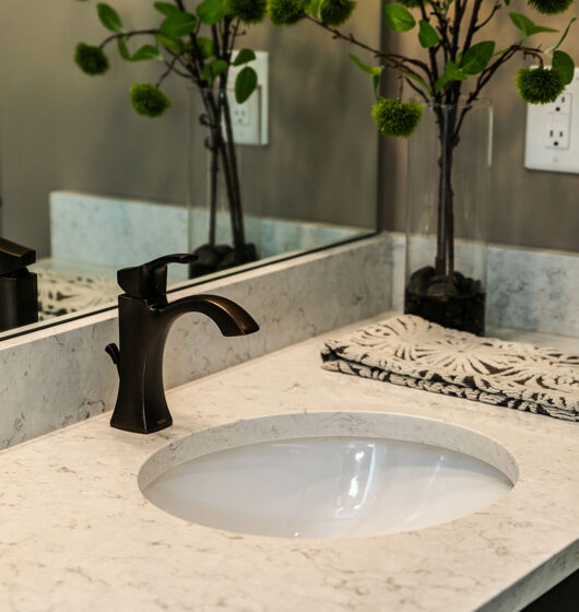 A bathroom sink with a bronze faucet on a marble countertop. A decorative towel with an abstract pattern lies beside it. Reflections of tall branches with green foliage are seen in the mirror behind the sink.