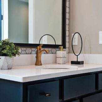 A modern bathroom sink with a white countertop, gold faucet, and dark cabinetry. On the counter are a potted plant, a circular mirror, and a soap dispenser. Hexagonal tiles form a decorative backsplash behind the sink.