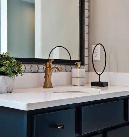 A modern bathroom sink with a white countertop, gold faucet, and dark cabinetry. On the counter are a potted plant, a circular mirror, and a soap dispenser. Hexagonal tiles form a decorative backsplash behind the sink.