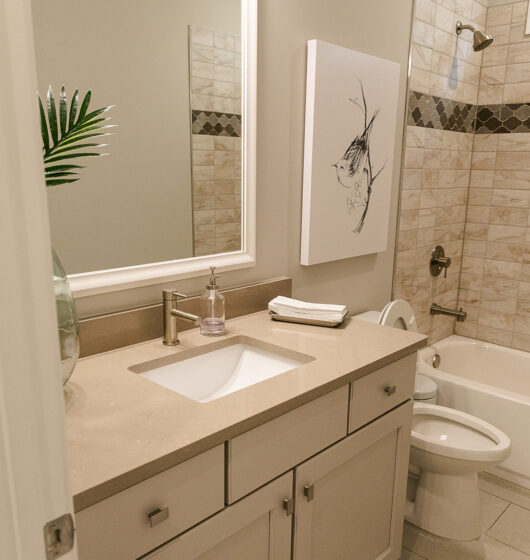 A modern bathroom with a beige vanity featuring a white sink and silver faucet. Above is a large mirror. A vase with a green leaf and folded towels sit on the countertop. The walls have tan tiles with decorative accents and a bird artwork hangs near the shower.