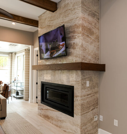 Modern living room with a stone fireplace and mounted TV. Dark wood beams accent the ceiling, complementing the wood mantel on the fireplace. Neutral tones dominate the room, with patterned carpeting and light-colored walls. Bright natural light streams in.