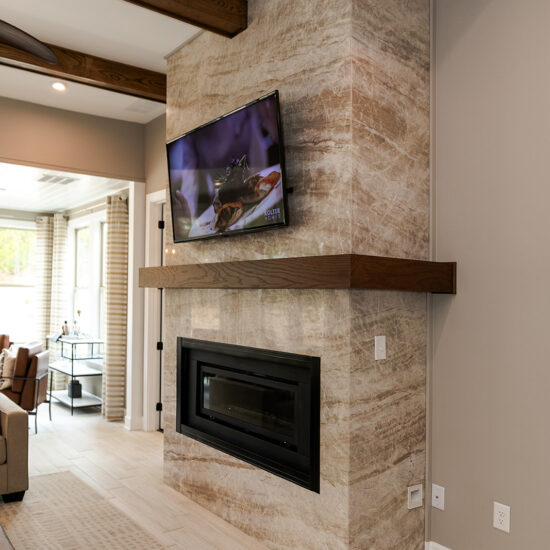 Modern living room with a stone fireplace and mounted TV. Dark wood beams accent the ceiling, complementing the wood mantel on the fireplace. Neutral tones dominate the room, with patterned carpeting and light-colored walls. Bright natural light streams in.