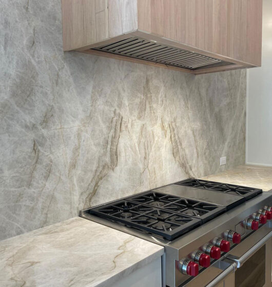 Modern kitchen with a light marbled backsplash and countertop, featuring a stainless steel gas stove with a range hood. The stove has red knobs and is set against light wood cabinetry and a white cabinet wall with multiple drawers on the right.