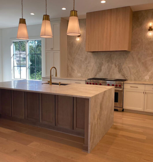 Modern kitchen with a large island featuring a beige countertop and wooden base, three pendant lights above, a stainless steel stove with a wooden hood, marble backsplash, white cabinets, and wooden flooring. Large windows allow natural light in.