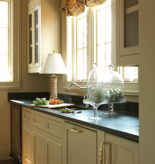 A sunlit kitchen counter with pale cabinets and a dark countertop. A table lamp, fruits, vegetables, and glass cloches with cupcakes are on the counter. Large windows with checkered curtains allow natural light to fill the space.