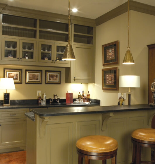 A cozy home bar with olive green cabinetry, dark countertops, and two brown leather stools. Three pendant lights hang above the bar. Framed artwork adorns the walls, and a wooden cabinet stands nearby. Bottles and glasses are displayed on the counter.