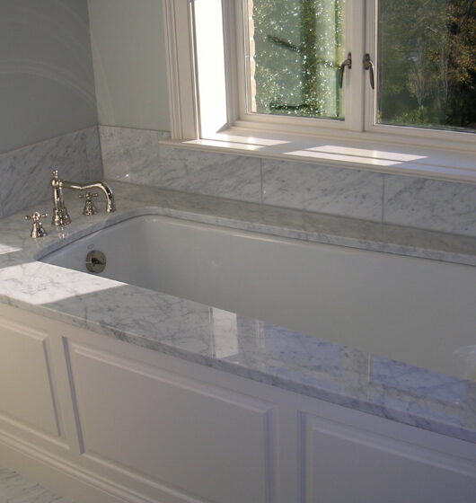 A white bathtub with marble trim is positioned under a window. The fixtures are silver, and the walls are light gray, complementing the marble detail. Sunlight streams through the window, creating slight reflections on the surface.