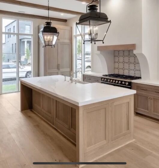 Modern kitchen with wooden floors and a large island featuring a white countertop. Two black pendant lights hang above the island. The room has wooden cabinets, a stove with a tile backsplash, and large windows letting in natural light.