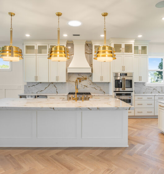 Modern kitchen with a large white marble island, gold accents, and three pendant lights. Cabinets are white with glass panels, and stainless steel appliances are built-in. The wood herringbone floor complements the sleek design.