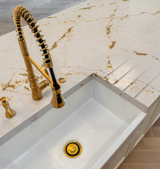 A modern kitchen sink with a tall black and gold faucet on a marble countertop with gold veining. The sink is rectangular and has a matching gold drain. The countertop features grooves for a draining board. Light wood flooring is visible below.