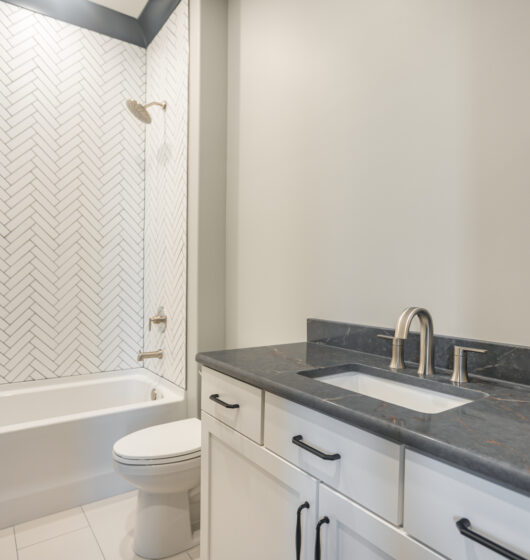 Modern bathroom with a white bathtub and toilet against a herringbone-patterned wall. The vanity has a dark marble countertop with a rectangular sink and silver faucet. White cabinets feature black handles, and the walls are light gray.