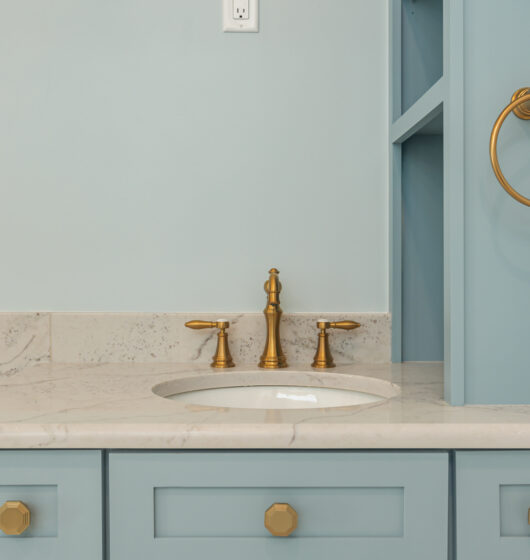 A bathroom sink with light blue cabinetry and a marble countertop. Gold-colored fixtures include the faucet, drawer handles, and a towel ring mounted on a nearby wall. The wall above is painted a pale blue.