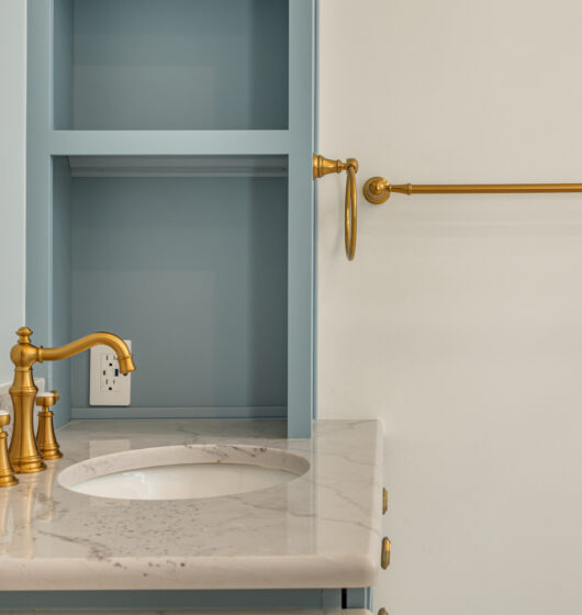 A bathroom vanity with a marble countertop and gold faucet set, set against a light blue wall. A gold towel bar is mounted on a white wall beside a built-in shelf. An electrical outlet is visible above the counter.