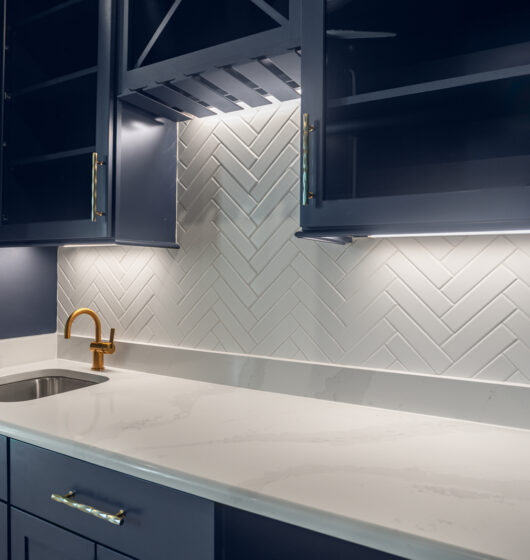 A modern kitchen with dark blue cabinets, a white marble countertop, and a gold faucet. The backsplash features white herringbone tiles, and there is under-cabinet lighting. The sink is small and sleek.