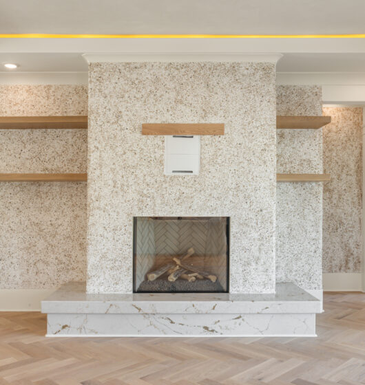 Luxurious living room with a stone fireplace, wooden shelves on either side, and a herringbone wood floor. Recessed lighting highlights the textured wall and sloped ceiling, creating a warm and inviting atmosphere.