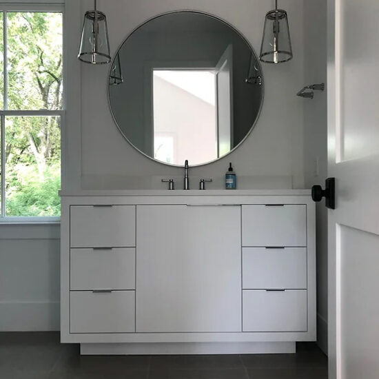 Modern bathroom with a white vanity featuring six drawers and a countertop. A round mirror is above the sink, flanked by two pendant lights. A window on the left shows greenery outside. Neutral-colored tiles cover the floor.